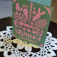Handmade Easter greeting card with bunny and floral cutouts in green and pink design, placed on a lace table mat.