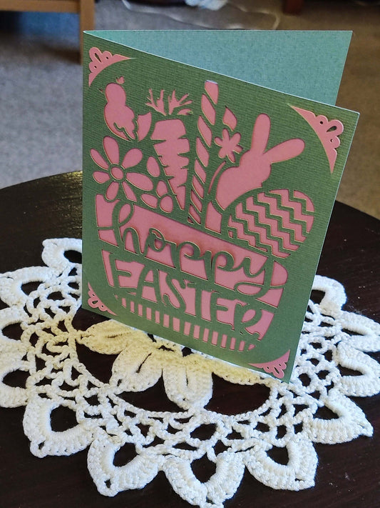 Handmade Easter greeting card with bunny and floral cutouts in green and pink design, placed on a lace table mat.