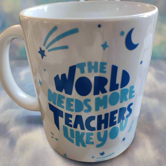 A white ceramic mug with blue text that reads: "THE WORLD NEEDS MORE TEACHERS LIKE YOU." Surrounding the text are blue decorative elements, including stars, a crescent moon, and abstract shapes resembling shooting stars. The mug has a handle on the left and rests on a neutral surface, radiating a sense of appreciation for educators.