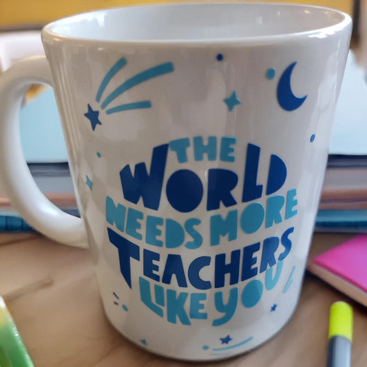 A white ceramic mug with a motivational message in blue text that reads, "THE WORLD NEEDS MORE TEACHERS LIKE YOU." The design includes decorative elements such as stars, a crescent moon, and abstract shapes. The mug is placed on a wooden table surrounded by stationery items like highlighters, pens, and a notebook, creating a cosy and inspiring workspace atmosphere.