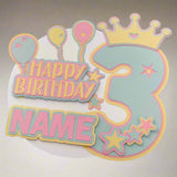 Personalised pastel 3rd birthday cake topper with balloons, stars, and crown, photographed from a direct top-down angle against a softly lit neutral background.