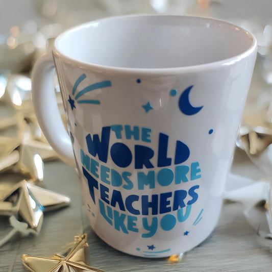 A white ceramic mug surrounded by golden star-shaped decorations. The mug features blue text and graphics, including stars, a crescent moon, and dynamic lines. The message on the mug reads: "THE WORLD NEEDS MORE TEACHERS LIKE YOU." The festive and inspiring design celebrates educators and their invaluable contributions.