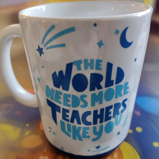 A white ceramic mug featuring blue text that reads, "THE WORLD NEEDS MORE TEACHERS LIKE YOU." The design is adorned with stars, a crescent moon, and shooting stars for a whimsical touch. The mug is displayed on a colourful surface with abstract patterns, creating a vibrant and uplifting presentation.