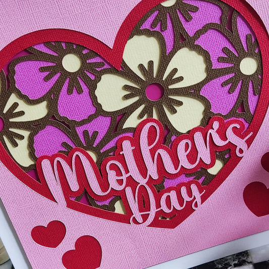 Handmade Mother's Day card with floral heart design in pink and purple, featuring cut-out flowers and layered hearts.