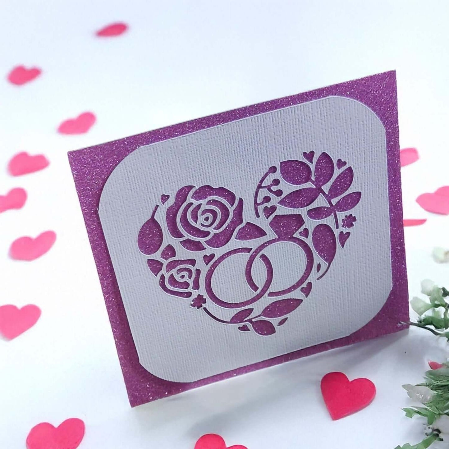 Handmade wedding or anniversary card with a sparkly purple background and a white die-cut floral heart design with wedding rings.