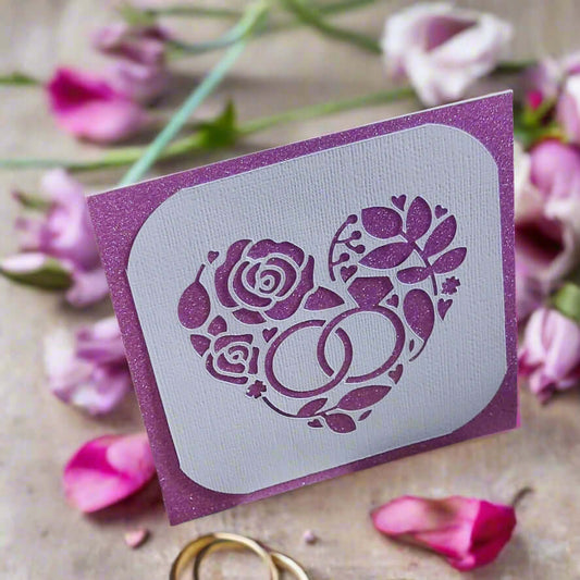 Handmade wedding or anniversary card with a sparkly purple background and a white die-cut floral heart design with wedding rings, displayed with actual wedding bands and pink petals.
