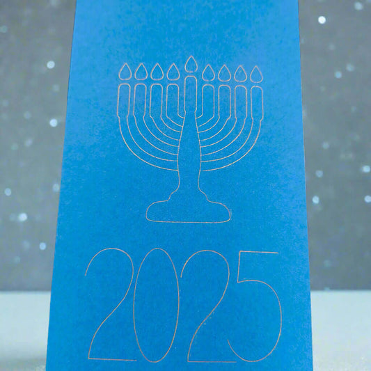 A bright blue Hanukkah card featuring a white line illustration of a menorah with nine candles above the year '2025'. The card stands upright against a soft grey background with scattered white dots resembling falling snow or festive lights, creating a peaceful and celebratory atmosphere.