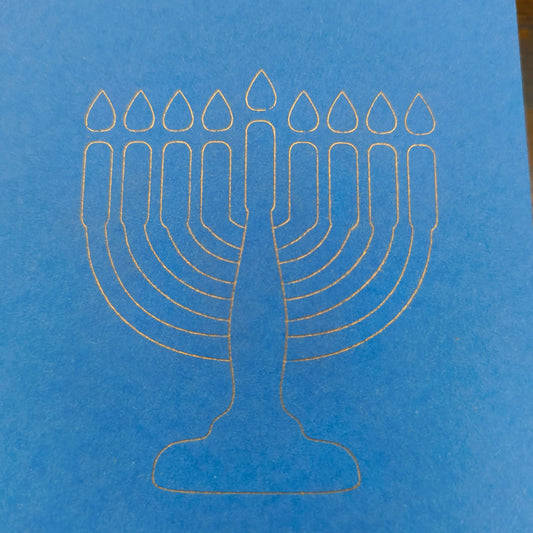 A close-up of a minimalist white line illustration of a menorah with nine candles, finely etched on bright blue card. The design is clean and modern, set against the smooth texture of the card, symbolising Hanukkah in a simple yet elegant style.