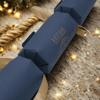 Navy blue personalised Christmas cracker with gold ribbon, displayed on a cosy fur rug with festive tree lights twinkling in the background.