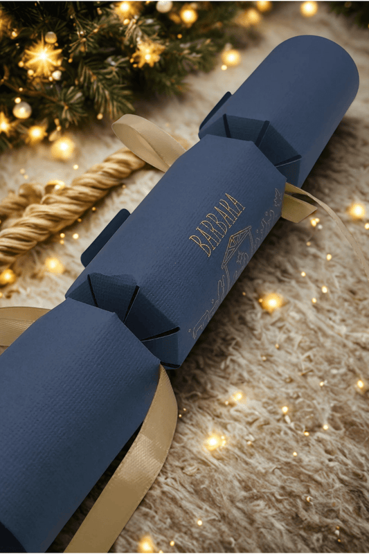 Navy blue personalised Christmas cracker with gold ribbon, displayed on a cosy fur rug with festive tree lights twinkling in the background.
