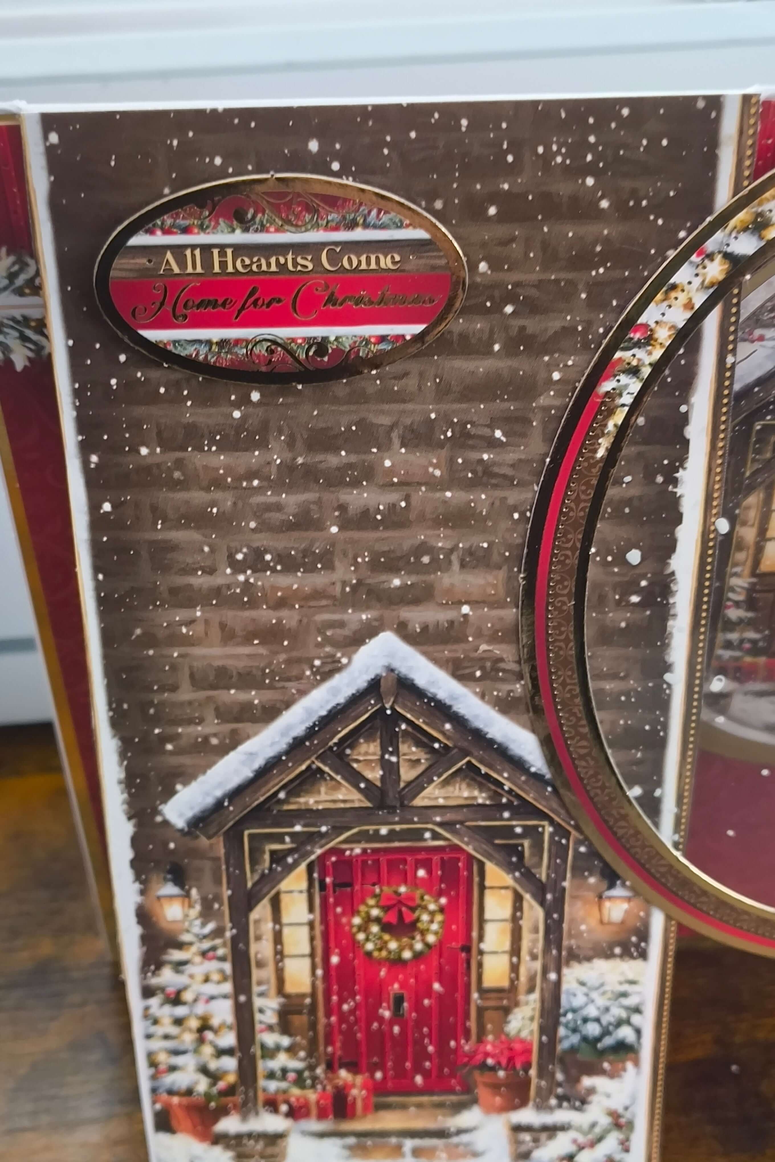 Close-up of Christmas card sentiment reading “All Hearts Come Home for Christmas” with gold edging and snowfall effect.