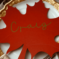 Autumn wedding place setting with a red maple leaf place card featuring the name Craig in gold script, displayed on a white plate with ornate gold trim and neutral linen napkin.