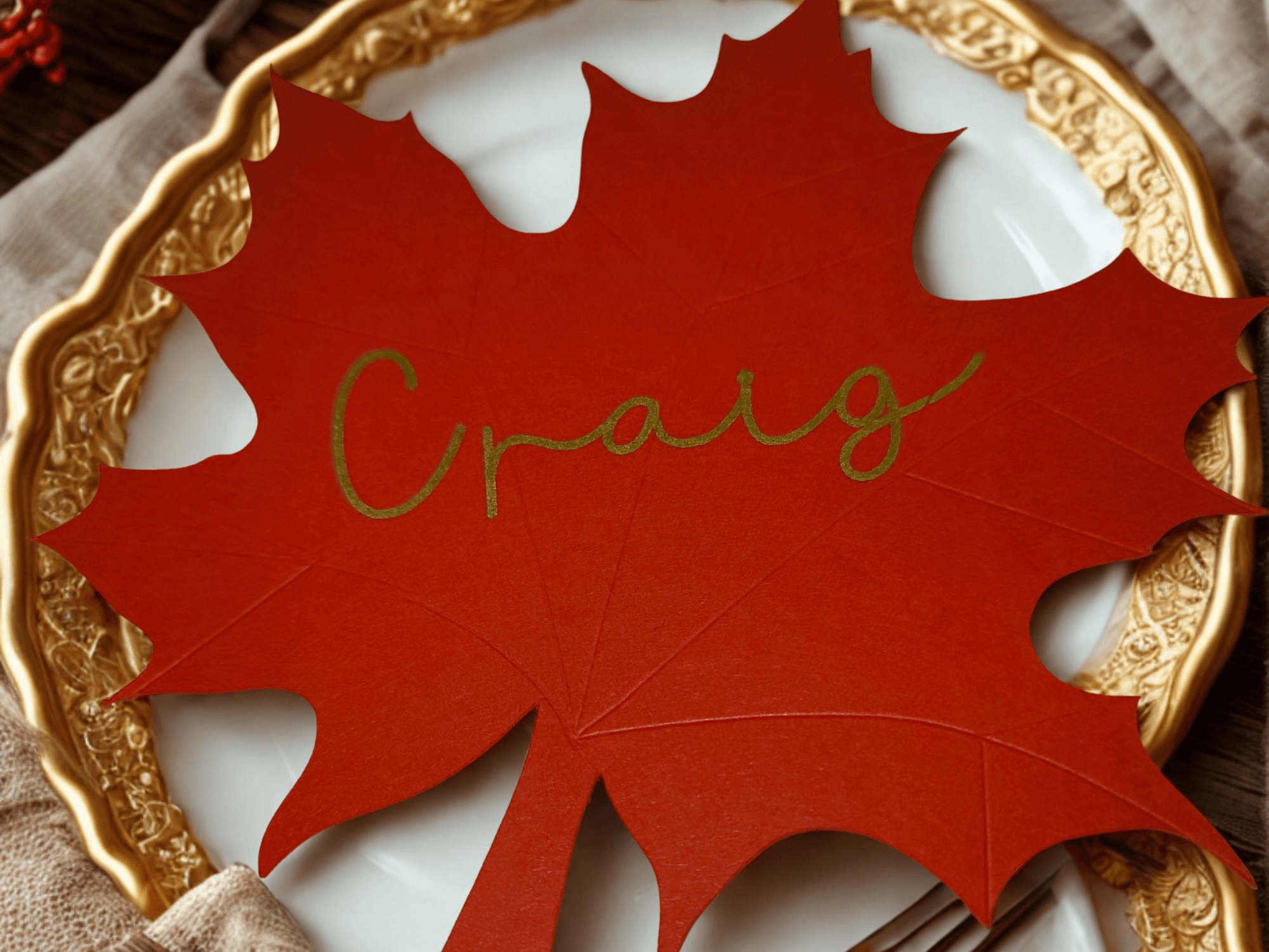 Autumn wedding place setting with a red maple leaf place card featuring the name Craig in gold script, displayed on a white plate with ornate gold trim and neutral linen napkin.