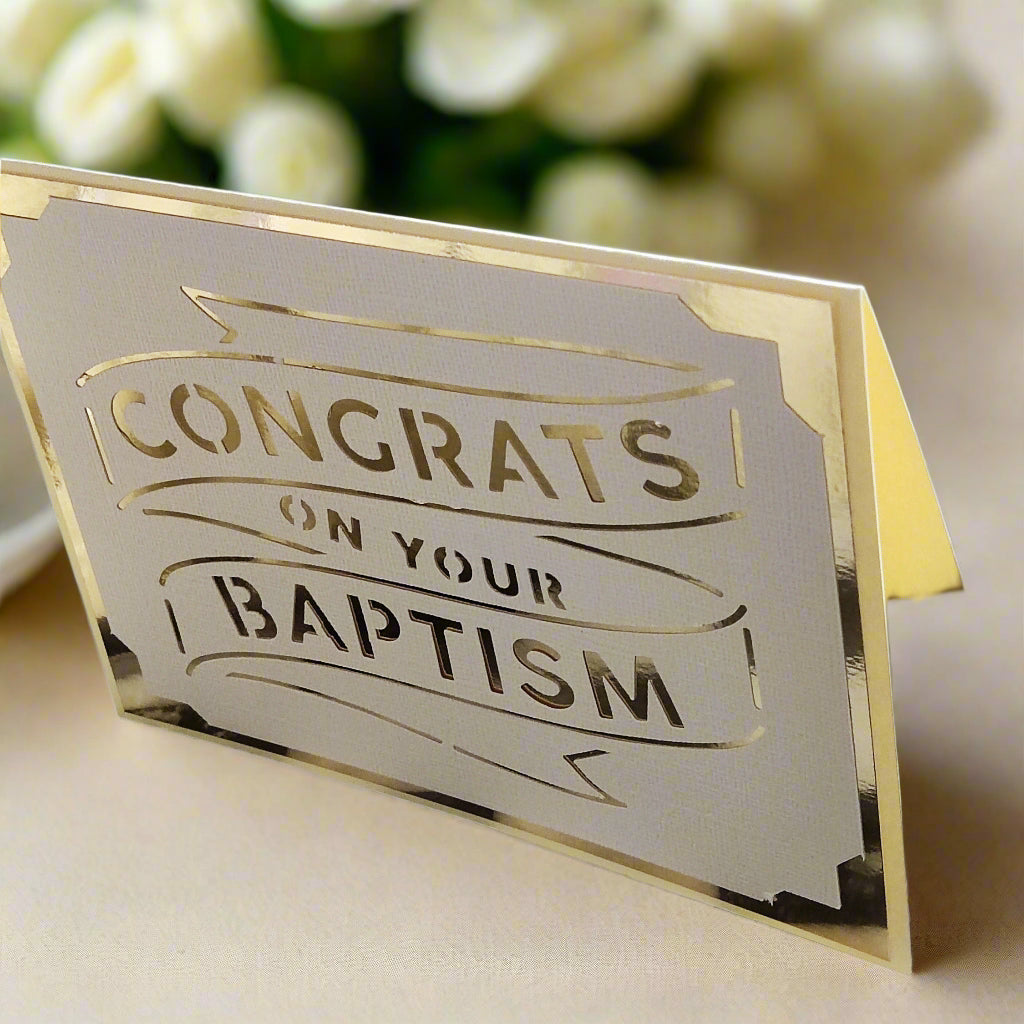 Elegant handmade baptism card with metallic gold foil cut-out text reading 'Congrats on Your Baptism', displayed on a soft cream surface with blurred white roses in the background.