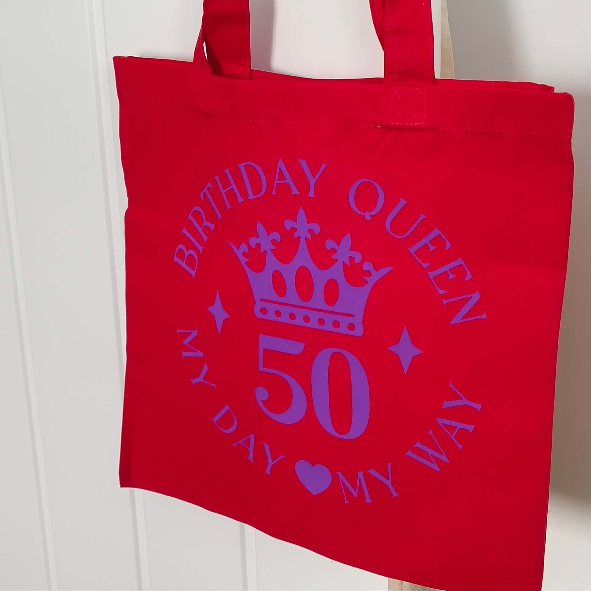 Red birthday queen tote bag with purple crown and 50th birthday design hanging neatly on door.