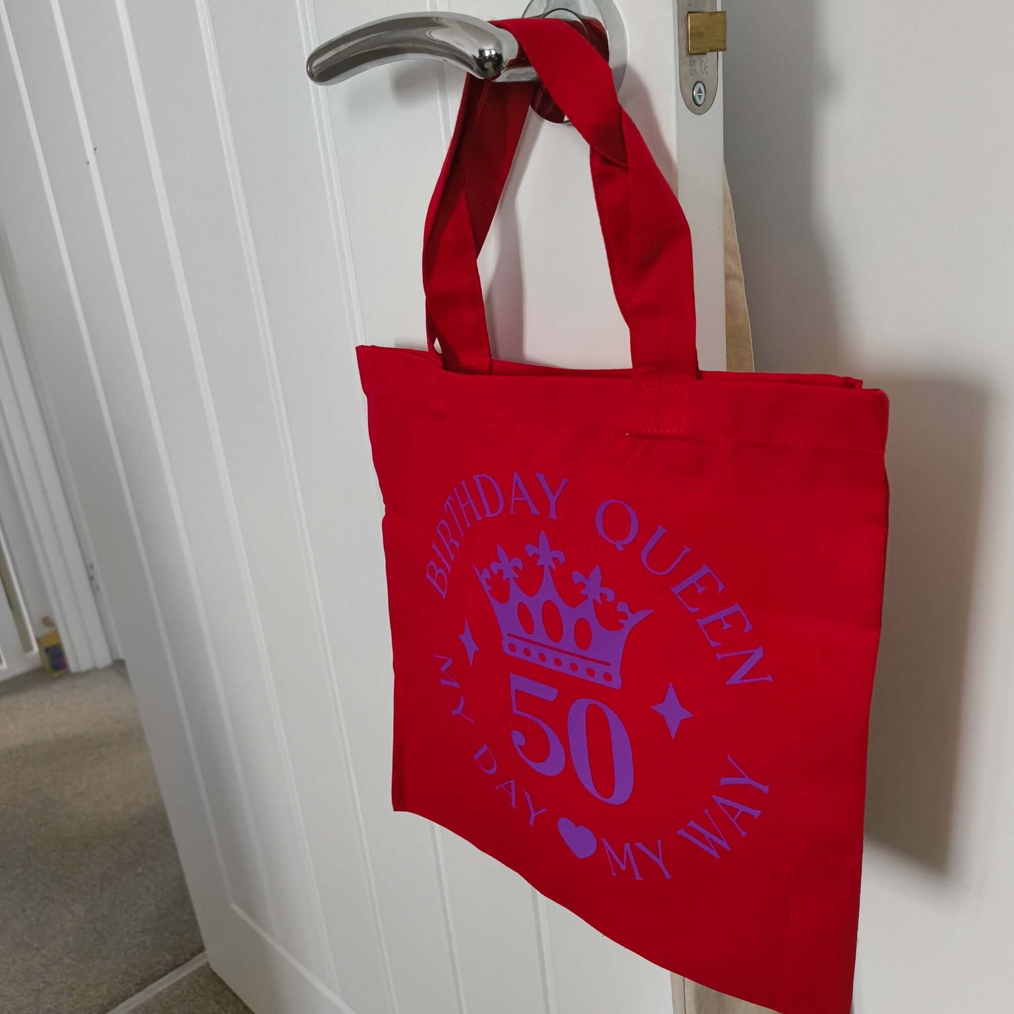 Red birthday queen tote bag hanging on the door handle, purple crown design with the number 50.