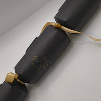 Black cracker tied with gold ribbon, showing a witch hat “Mum” design on the centre panel