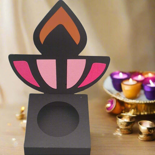 Front-facing image of a black Diwali sweet holder with pink and orange diya design, displayed on a wooden table with festive tealights and gold accents in the background.