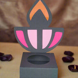 Front-facing image of a handmade black Diwali diya sweet holder with pink and orange flame design, displayed on a beige surface with rose petals and soft festive lighting in the background.