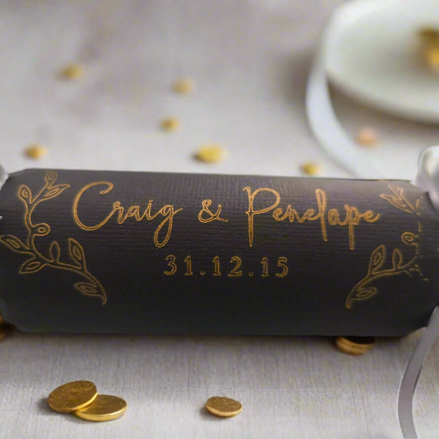 Black wedding cracker with gold foil personalisation, featuring names and date, tied with silver ribbons, styled with metallic gold leaves on a grey background.