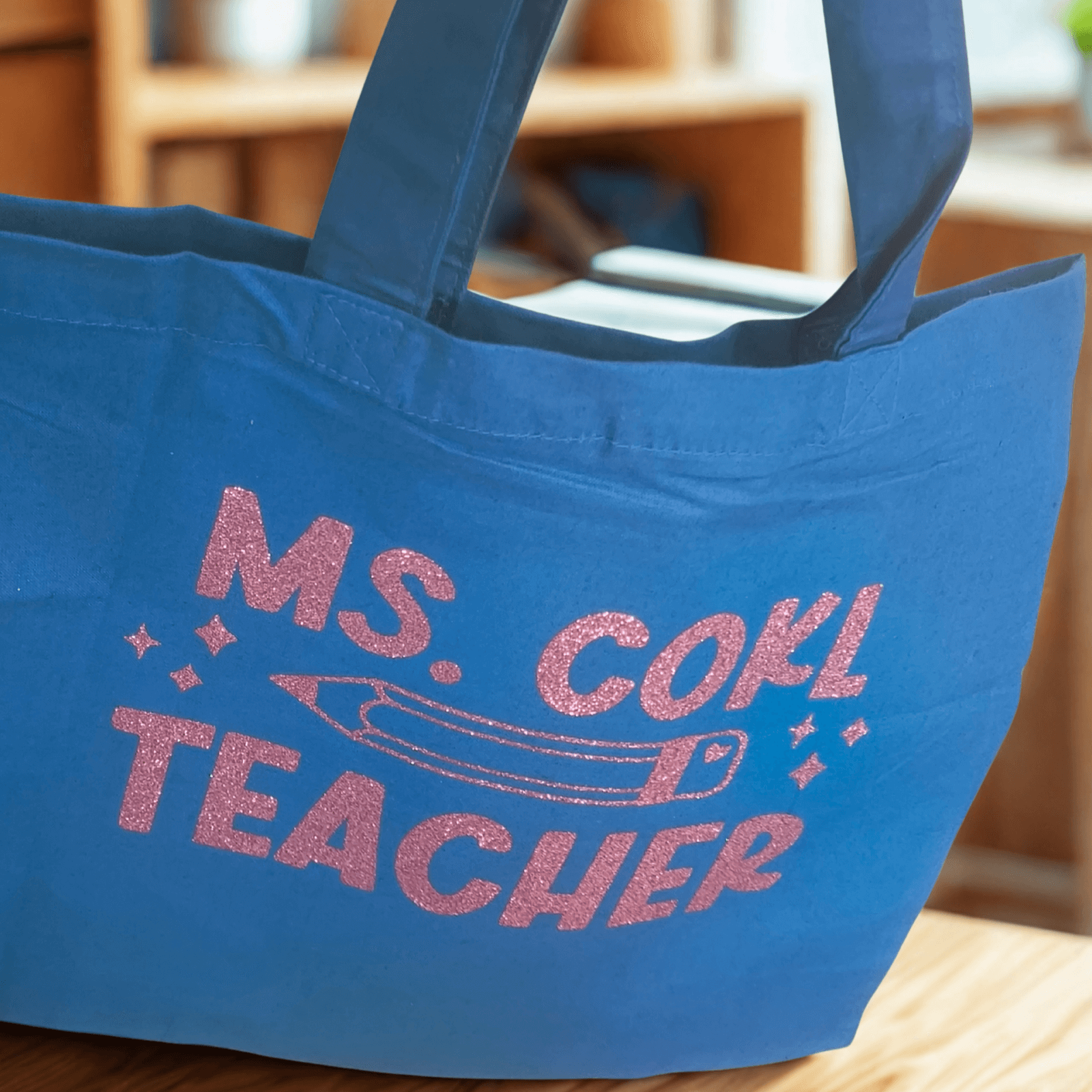 Blue teacher tote bag with pink glitter vinyl text "Ms. Cohl Teacher" and pencil design, hanging on a door handle.