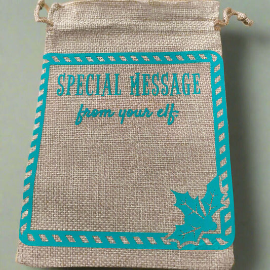 Burlap drawstring gift bag with turquoise vinyl text reading 'Special Message from your elf', framed with a candy cane-style border and a holly leaf design in the bottom right corner.