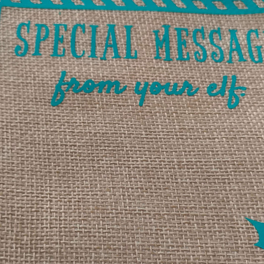 Partial close-up of a burlap gift bag with turquoise vinyl text reading 'Special Message from your elf', showing the woven fabric texture and decorative border.