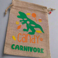 Handmade hessian drawstring bag with a green dinosaur graphic surrounded by colourful sweets and the phrase 'Candy Carnivore' in orange and green text. The bag is displayed on a plain light grey background.