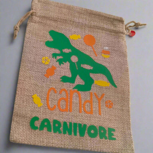 Handmade hessian drawstring bag with a green dinosaur graphic surrounded by colourful sweets and the phrase 'Candy Carnivore' in orange and green text. The bag is displayed on a plain light grey background.