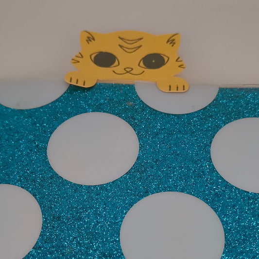 Handmade yellow cat paper cut-out peeking over the edge of a blue glittery polka dot surface, resembling a bookmark.