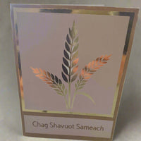 A handmade "Chag Shavuot Sameach" greeting card featuring a metallic gold and copper wheat design, framed with a gold foil border, displayed against a soft, light beige fabric background.