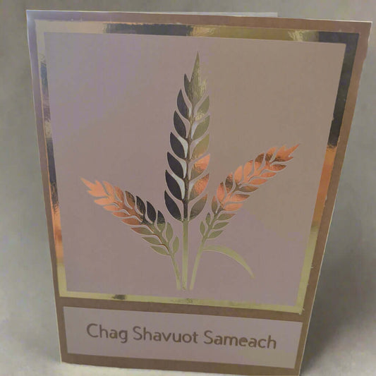 A handmade "Chag Shavuot Sameach" greeting card featuring a metallic gold and copper wheat design, framed with a gold foil border, displayed against a soft, light beige fabric background.