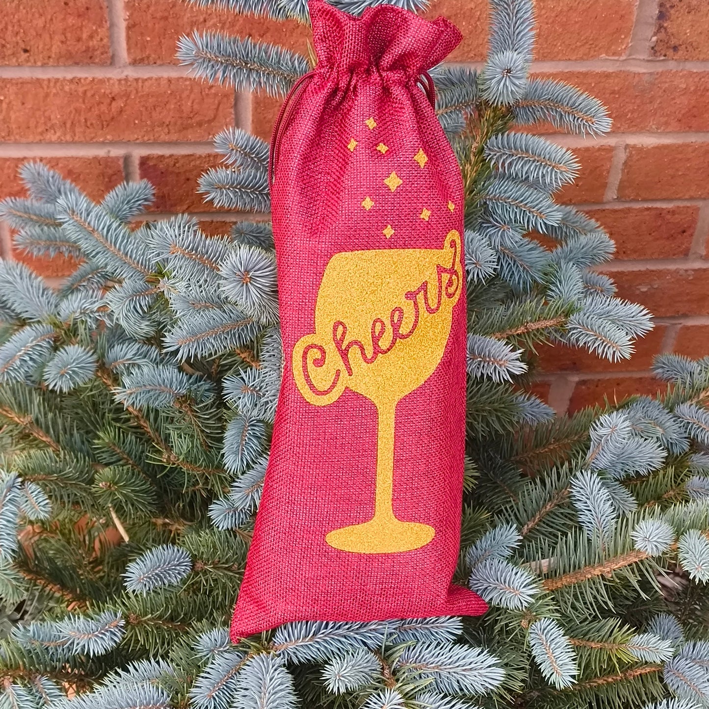 Red wine bottle gift bag with gold glitter wine glass and cheers text against brick wall background.