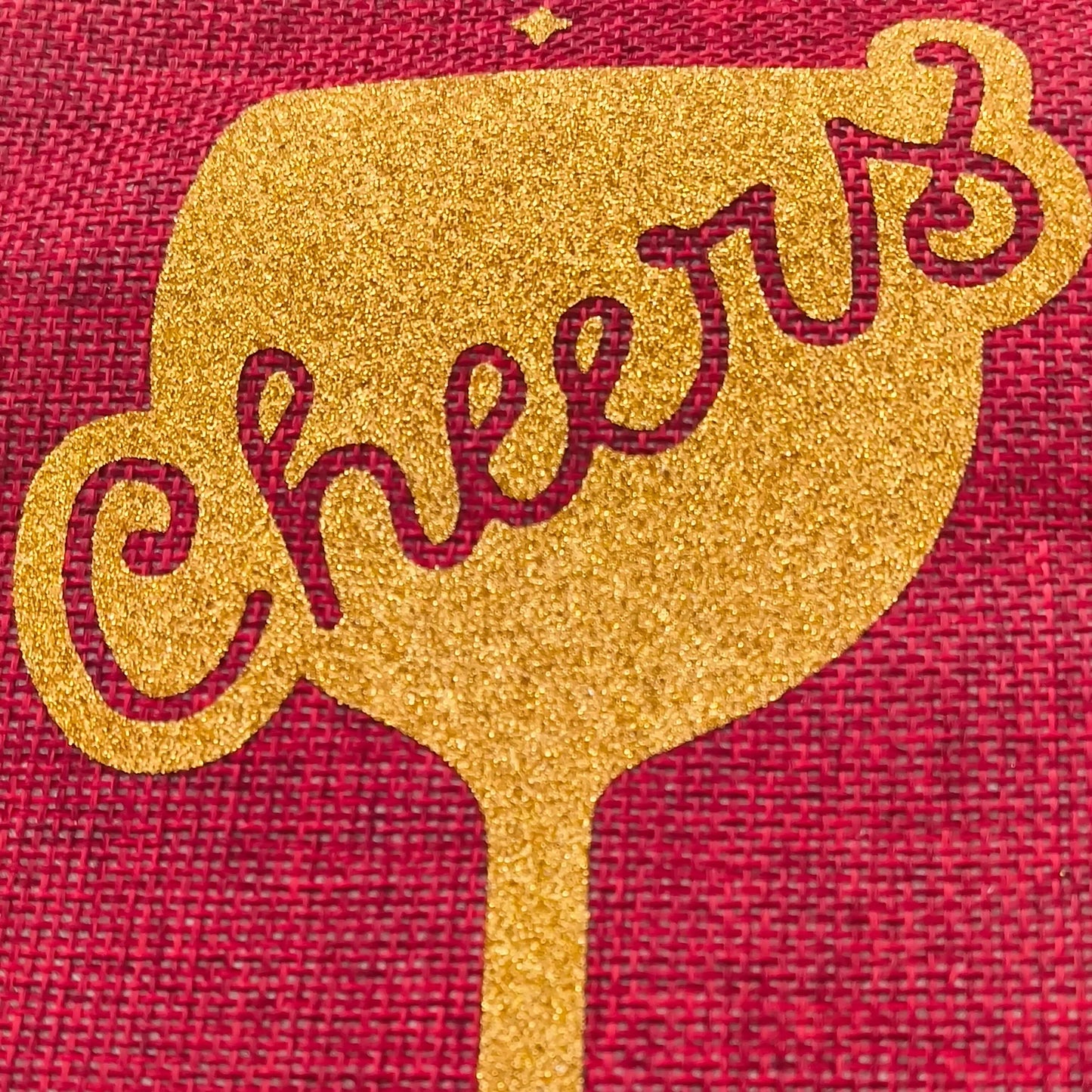 Extreme close-up of gold glitter 'Cheers' wine glass design on a red hessian gift bag, highlighting sparkle texture and woven fabric detail.