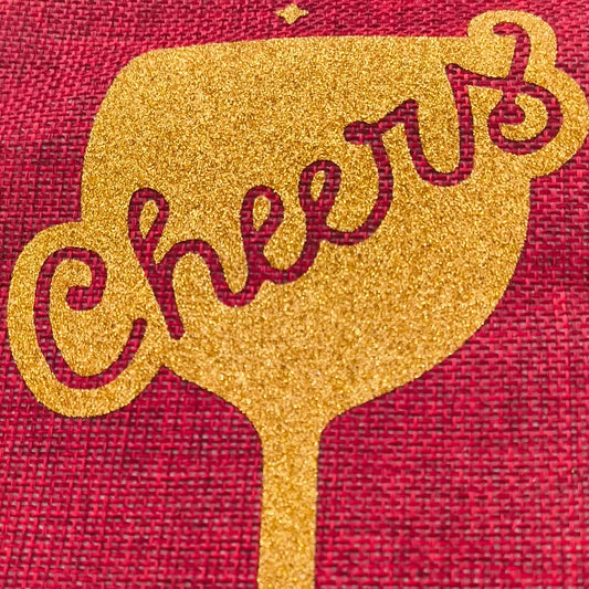 Extreme close-up of gold glitter 'Cheers' wine glass design on a red hessian gift bag, highlighting sparkle texture and woven fabric detail.