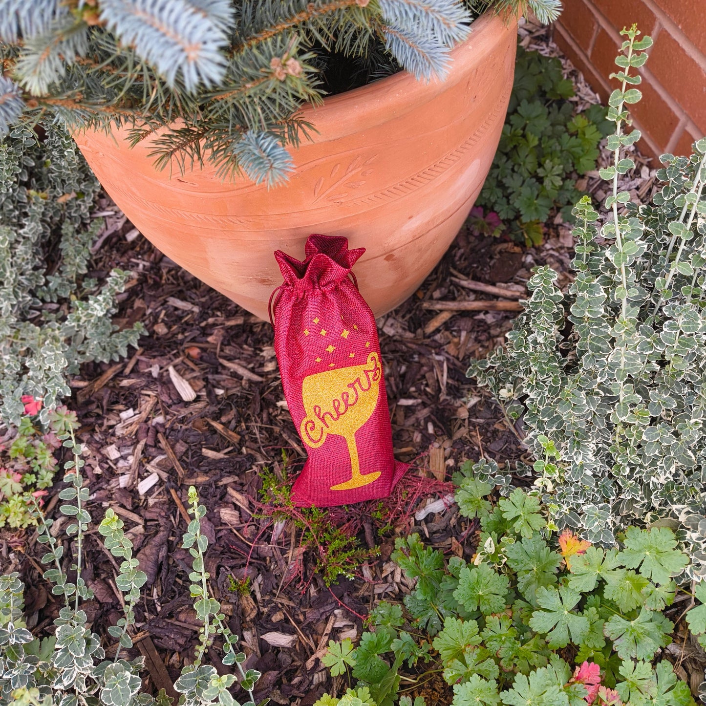 Red reusable wine bag with gold glitter cheers design resting at base of plant pot.