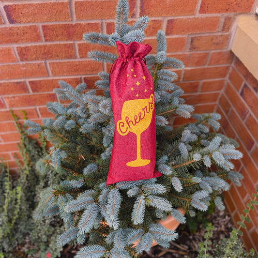 Cheers wine gift bag in red hessian with gold glitter design shown outdoors in garden setting.