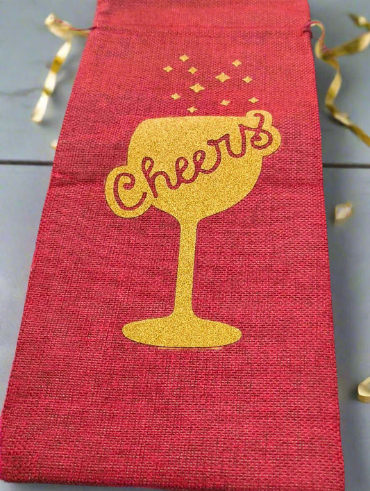 Red wine bottle gift bag with gold glitter 'Cheers' design, photographed from a top-down angle on a grey background with curling gold ribbon for decoration.