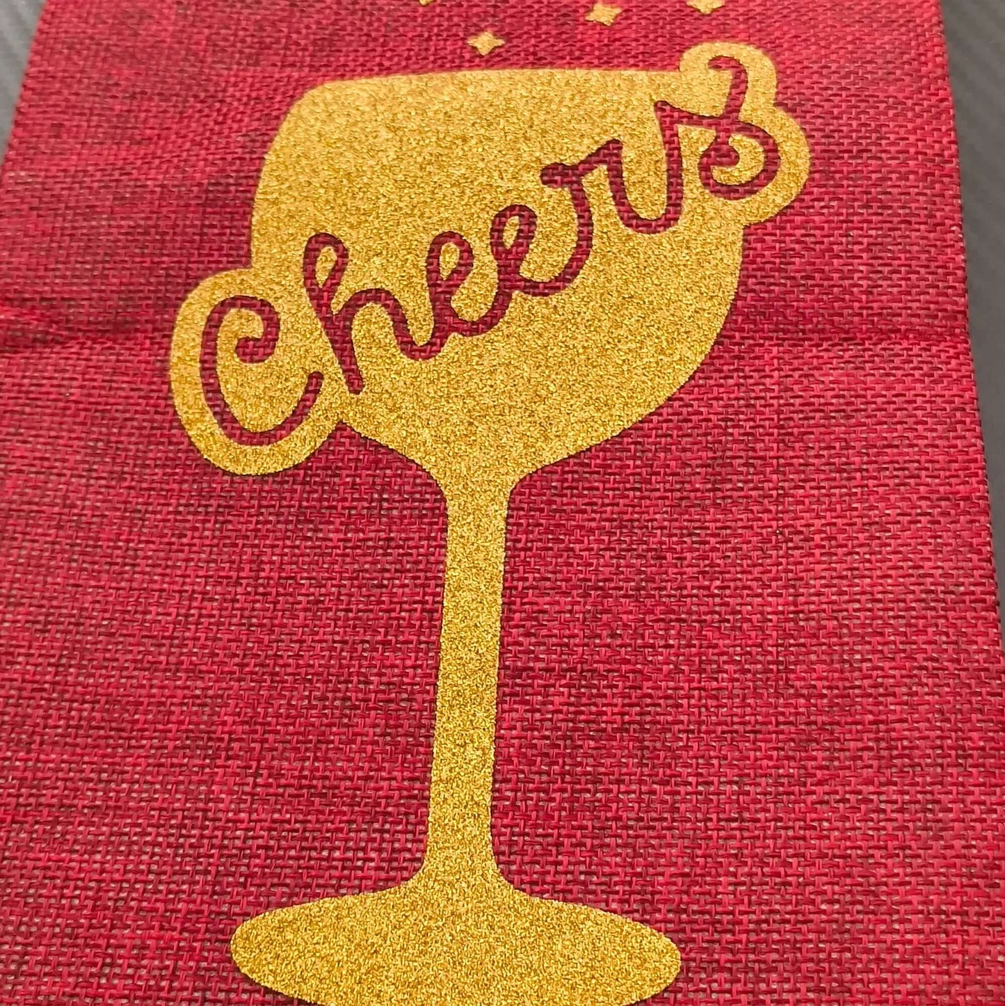 Close-up of a red hessian wine bottle bag featuring a gold glitter 'Cheers' wine glass design, viewed from a front-on angle, showcasing the texture and sparkle detail.