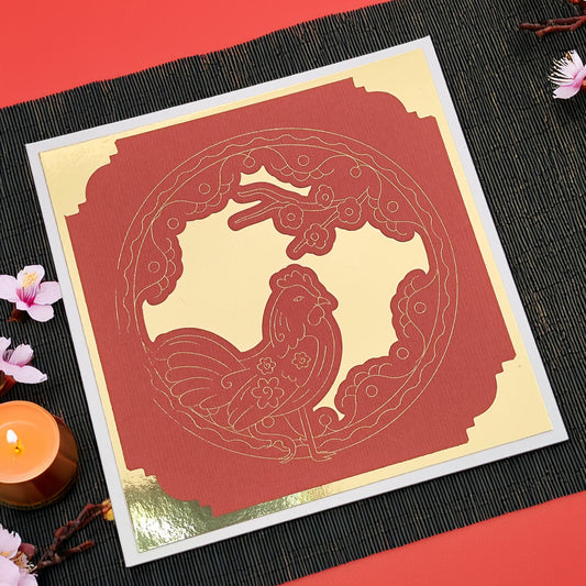 Flat lay of a red and gold Chinese New Year rooster greeting card on a black placemat, handmade with layered cardstock, perfect for Lunar New Year celebrations.