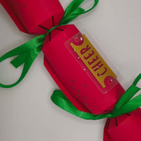 Christmas cracker in red with gold ‘CHEER’ tag and green satin ribbon, shown from above.
