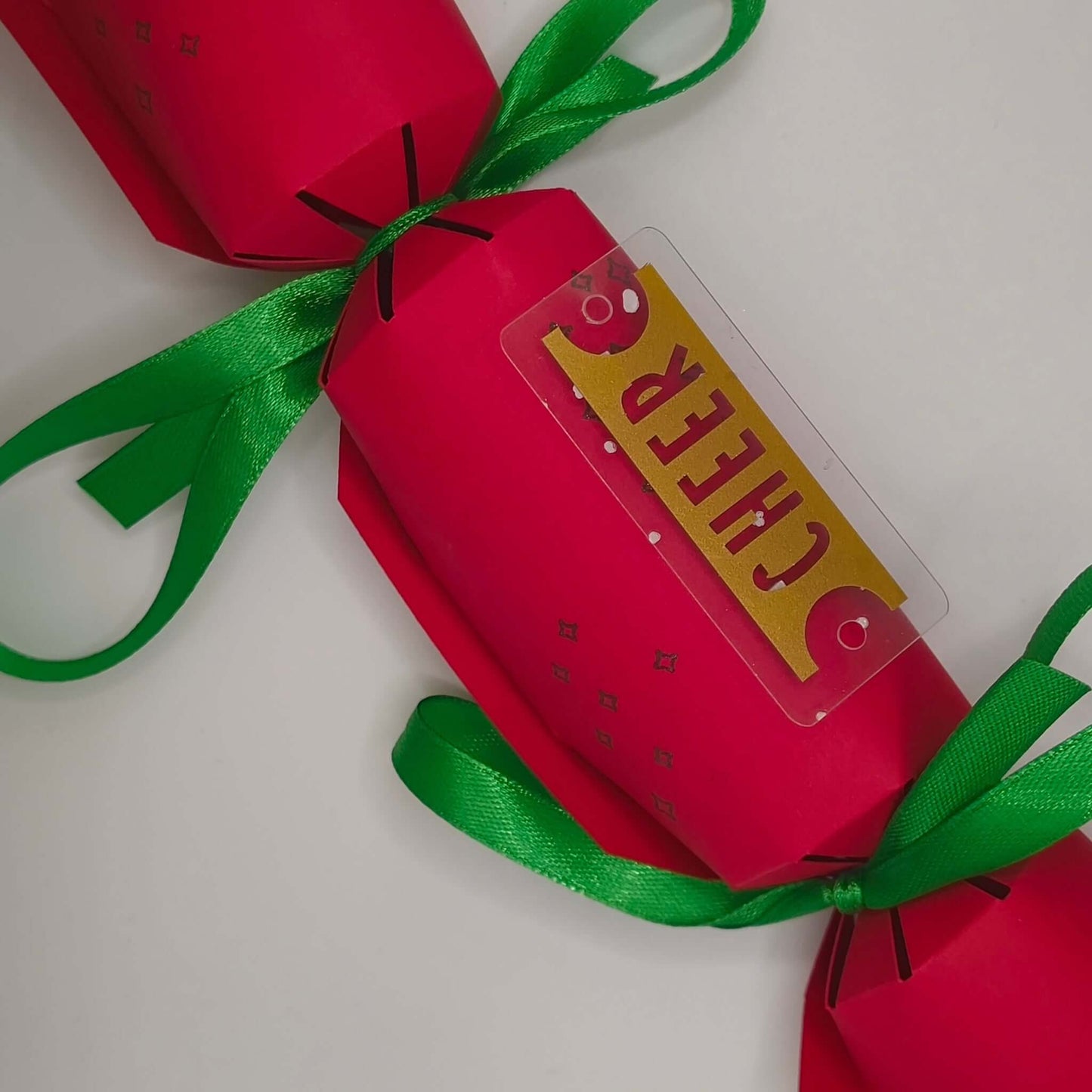 Christmas cracker in red with gold ‘CHEER’ tag and green satin ribbon, shown from above.