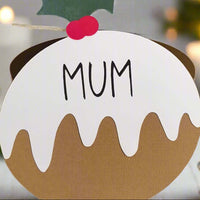 Close-up of Christmas pudding place card with “Mum” text in black lettering, white icing design, and holly topper with red berries, shown against a blurred festive background.