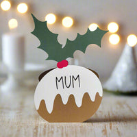 Christmas pudding-shaped place card with “Mum” written on it, decorated with green holly leaves and red berries, standing on a rustic wooden table with festive lights in the background.