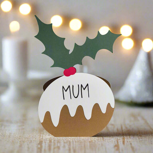 Christmas pudding-shaped place card with “Mum” written on it, decorated with green holly leaves and red berries, standing on a rustic wooden table with festive lights in the background.
