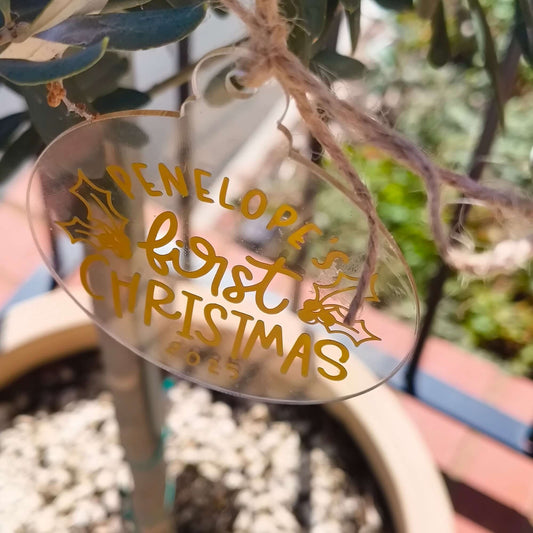 Personalised baby’s first Christmas bauble in clear acrylic with gold text hanging on tree.