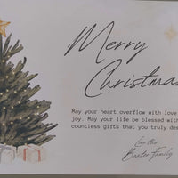 Close-up of a luxury handmade Christmas card featuring a decorated tree with a gold star topper and glowing lights, surrounded by wrapped presents. The card bears the message “Merry Christmas” in elegant script, with a heartfelt sentiment printed below and signed “Love the Baxter Family.” The design is bordered with gold and green details.