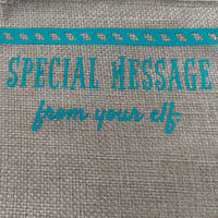 Close-up of turquoise vinyl text on a burlap surface reading 'Special Message from your elf', with part of a striped decorative border visible at the top