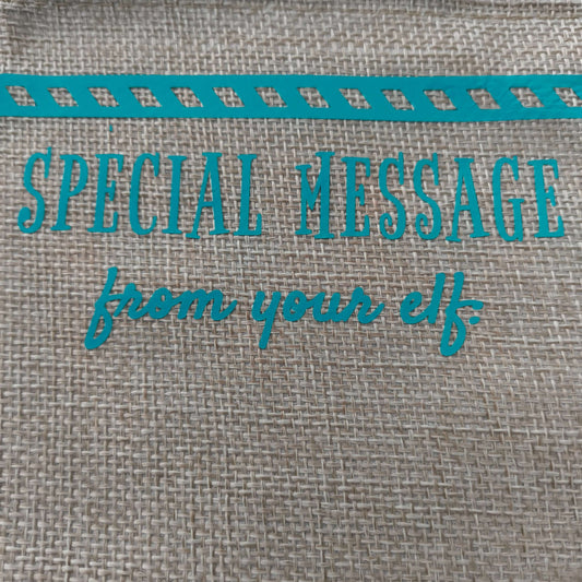 Close-up of turquoise vinyl text on a burlap surface reading 'Special Message from your elf', with part of a striped decorative border visible at the top