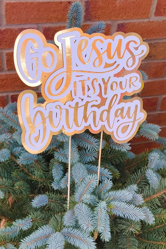 Close-up of a gold and white “Go Jesus It’s Your Birthday” cake topper with script lettering on a spruce tree.